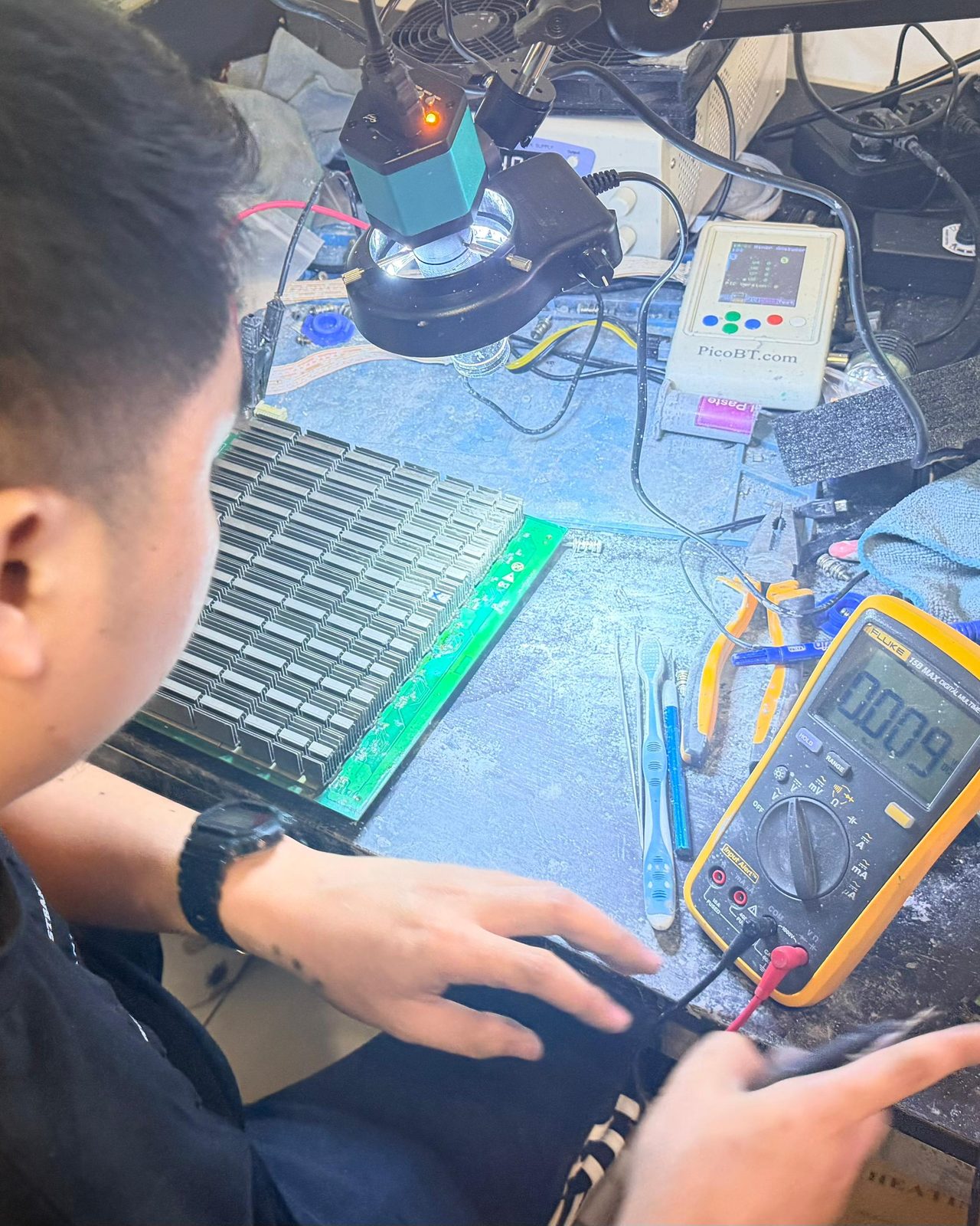 A hashboard with its heatsinks lying on the workshop bench, a hand and a multimeter beside it